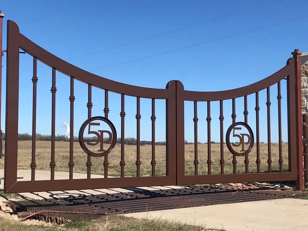 The 5P Gate closed across a ranch entrance, its bronze leaves dipping toward the centre, each carrying a 5P monogram in a rope-twist circle, with open pasture behind.