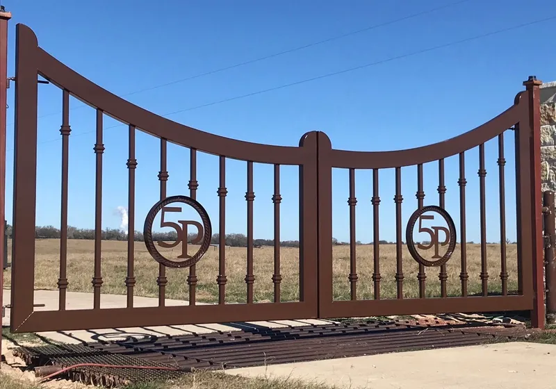 The 5P Gate closed across a ranch entrance, its bronze leaves dipping toward the centre, each carrying a 5P monogram in a rope-twist circle, with open pasture behind.