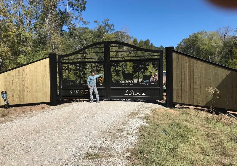 The Swazye Gate closed across a gravel drive between runs of black-capped wood privacy fence, its cut deer scene dark against green woods, with a man standing at the left leaf for scale.