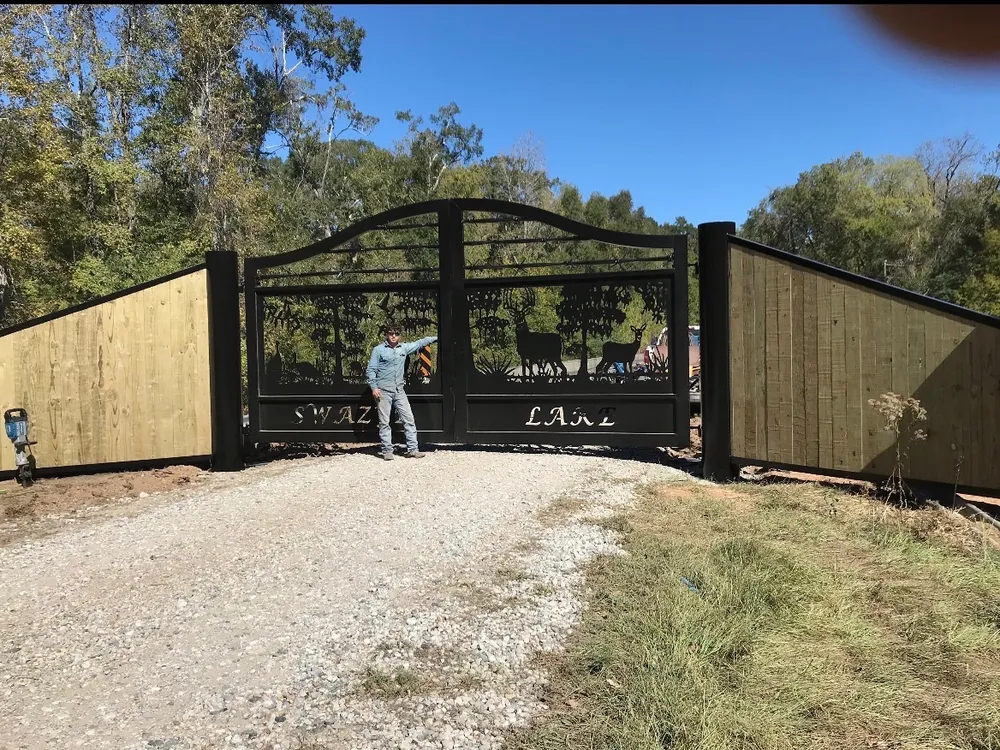 The Swazye Gate closed across a gravel drive between runs of black-capped wood privacy fence, its cut deer scene dark against green woods, with a man standing at the left leaf for scale.