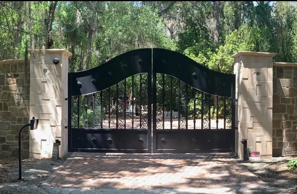 The Tampa Gate square-on and closed, centred between its two stone columns with the curved stone wall running off each side and Spanish moss overhead.