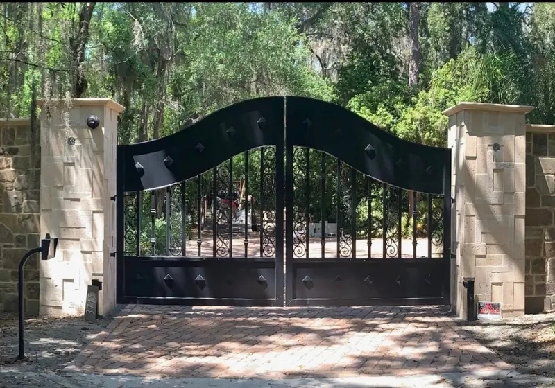 The Tampa Gate square-on and closed, centred between its two stone columns with the curved stone wall running off each side and Spanish moss overhead.