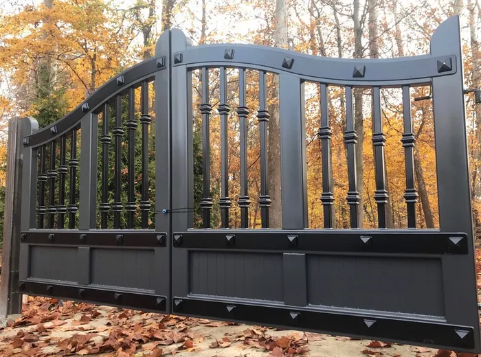 A dark grey build of the Ella Grace Gates on a leaf-strewn drive, turned balusters above a boarded lower panel banded with pyramid studs, autumn woodland behind.