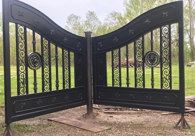 Both leaves of the New Yorker Gate hung on a bare steel post in a grassy yard and angled toward the camera, their gloss black rosettes, scrollwork and lion's-head rings bright under an overcast sky.