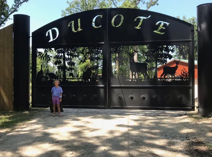 The gate square-on in full sun, a name cut through the arched top panel above a pierced scene of deer, turkeys and timber, with a child standing at the left leaf for scale.
