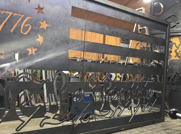 The gate in bare steel inside the shop, hanging from a chain hoist with the stars, date and lettering cut but unpainted, tool racks and a second cut panel behind it.