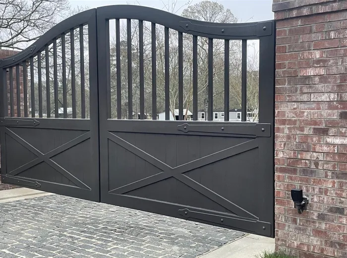 The Stable Gate closed up against a brick pillar on a cobblestone drive, its arched top rail and pickets above a boarded panel crossed by an X brace, strap plates and bolt heads marking the joints.