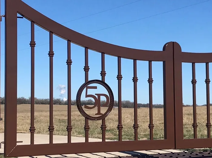 The left leaf of the 5P Gate close up, its monogram inside a rope-twist ring hung among collared pickets, with dry pasture and a treeline beyond.
