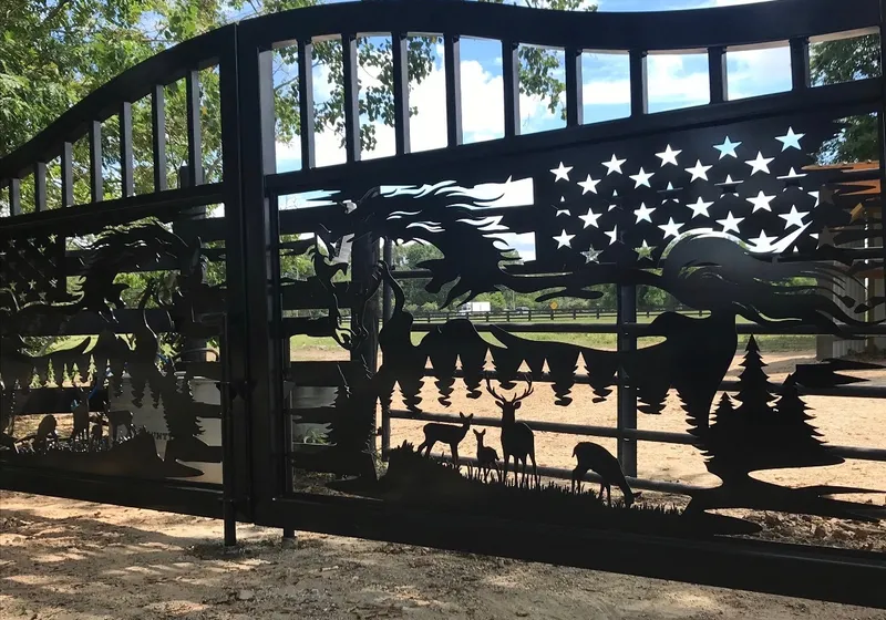 The Freedom Ranch Gate closed across a dirt drive and seen from one side, the near leaf's cut-out horse, flag and deer filling the frame with pasture, rail fencing and a cloudy sky showing through the openings.