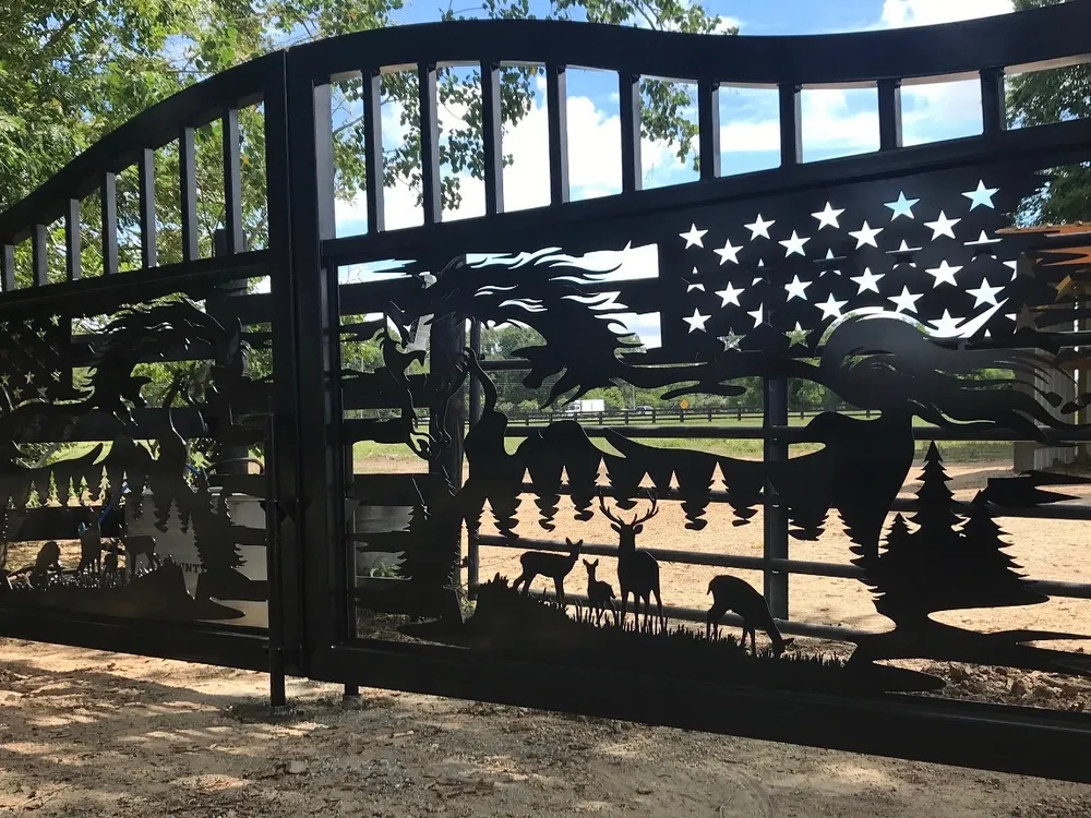 The Freedom Ranch Gate closed across a dirt drive and seen from one side, the near leaf's cut-out horse, flag and deer filling the frame with pasture, rail fencing and a cloudy sky showing through the openings.