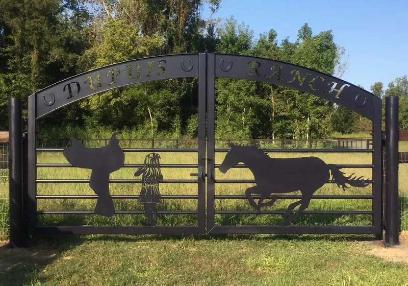 The Ranch Gate closed square-on across a grass drive, its arched band of cut lettering spaced by raised horseshoes above silhouettes of a saddle, a child and a galloping horse, with mown pasture and a treeline behind.