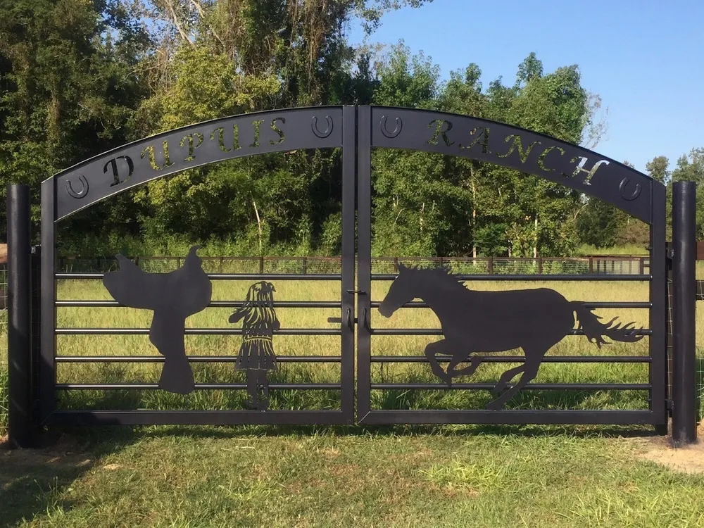 The Ranch Gate closed square-on across a grass drive, its arched band of cut lettering spaced by raised horseshoes above silhouettes of a saddle, a child and a galloping horse, with mown pasture and a treeline behind.