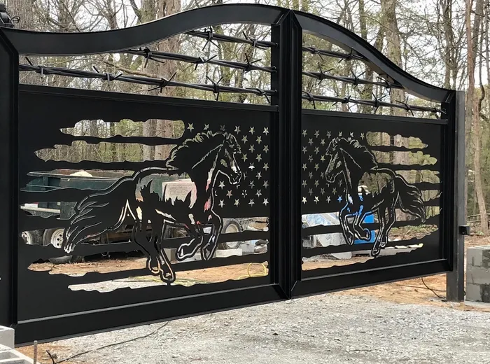 A second Frontier Gate on a gravel drive in the woods, galloping horses cut into the flag panel and barbed wire strung beneath the swept top rail.