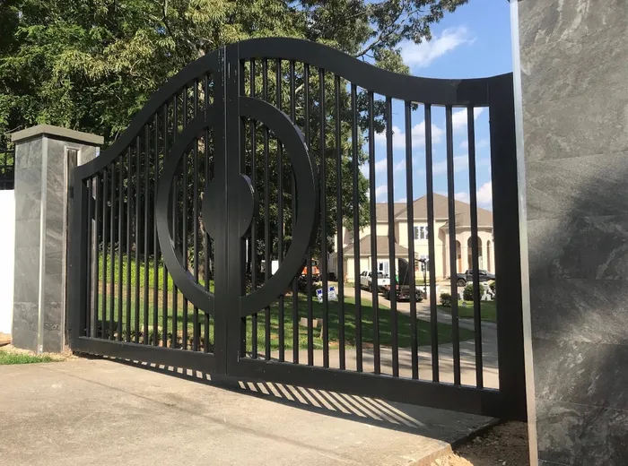 The Lake Spivey Gate seen along its length, the swept top rail cresting mid-span and running down to the far post, the ring showing its depth against the drive, with a pale arcaded house on the lawn behind.