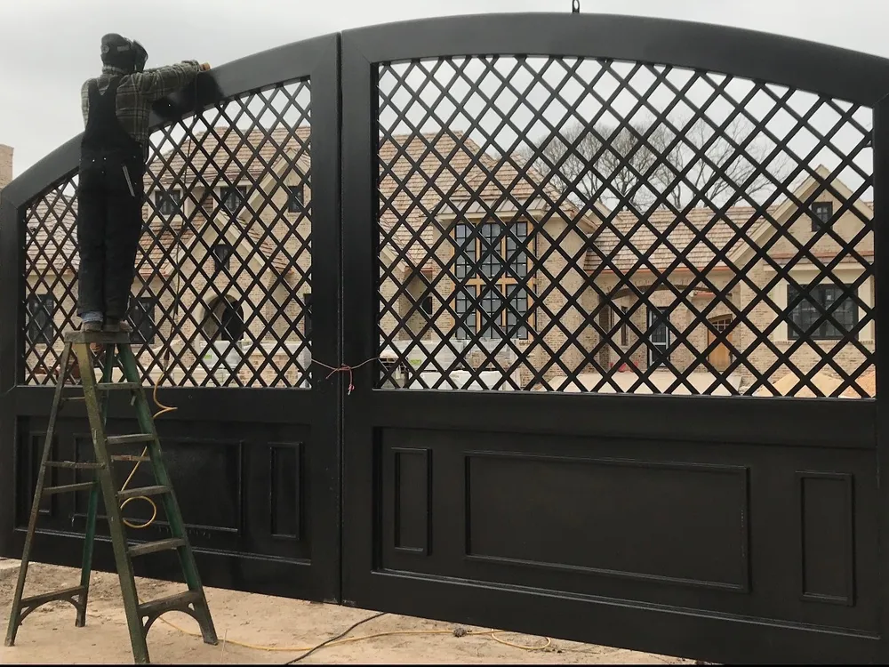 Both leaves of the Grand Lattice Gate hung and finished black at a stone house, the diamond lattice above raised lower panels, with a man in a welding hood on a step ladder working at the left leaf.