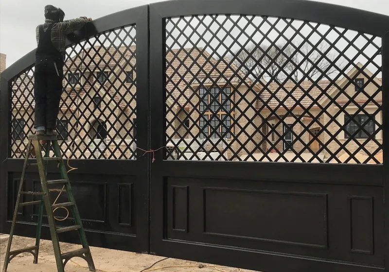 Both leaves of the Grand Lattice Gate hung and finished black at a stone house, the diamond lattice above raised lower panels, with a man in a welding hood on a step ladder working at the left leaf.
