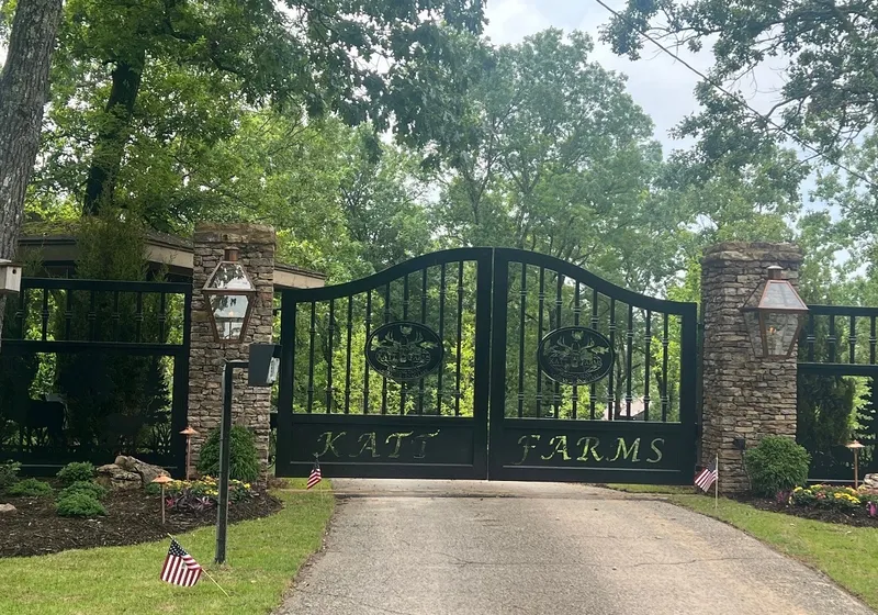 The Katt Gate closed across an asphalt drive between stone piers carrying copper gas lanterns on their faces, its oval medallions and lettered bottom panel dark against heavy summer trees, with matching picket fence panels running off to each side.
