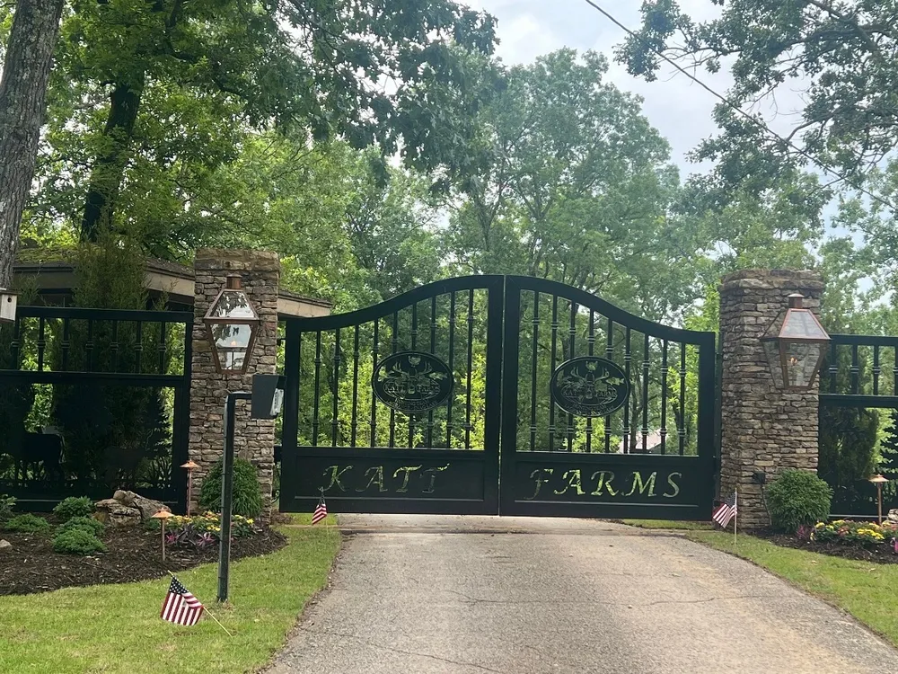 The Katt Gate closed across an asphalt drive between stone piers carrying copper gas lanterns on their faces, its oval medallions and lettered bottom panel dark against heavy summer trees, with matching picket fence panels running off to each side.