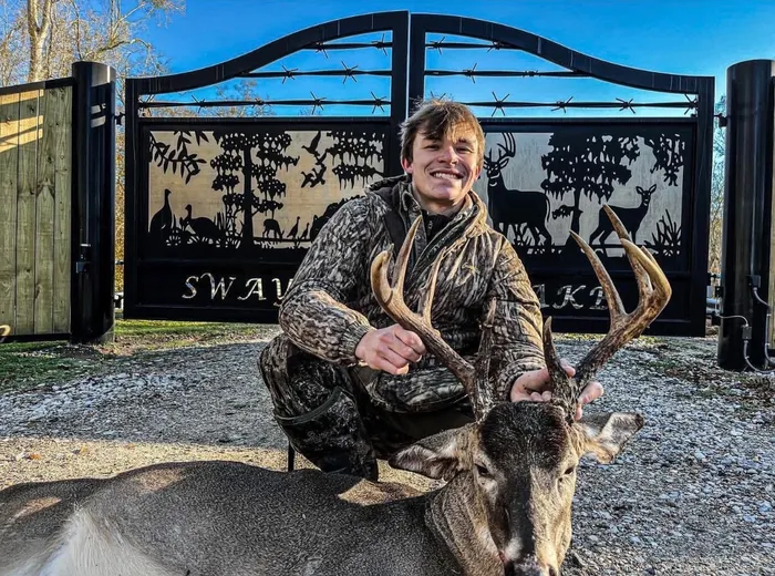 A hunter kneeling on the gravel in front of the closed gate, holding the antlers of a whitetail buck, with the cut woodland panels and barbed-wire top rails behind him.