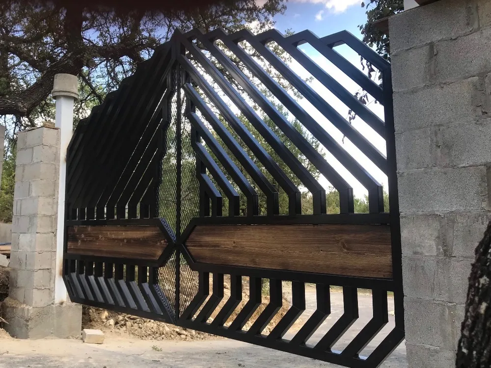 The Blackthorn Gate seen from the side of a drive, its stepped diagonal bars rising against a bright sky between unrendered concrete block columns, with a band of dark timber across the middle and evergreens beyond.
