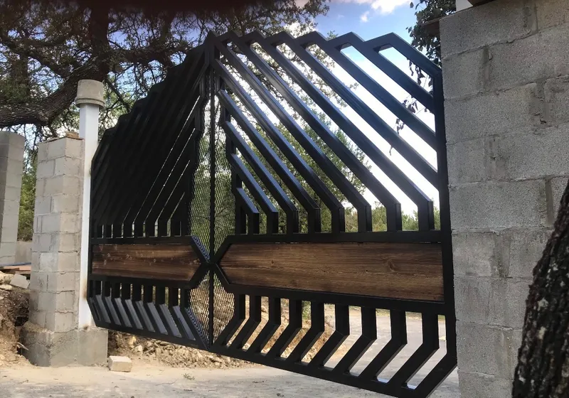 The Blackthorn Gate seen from the side of a drive, its stepped diagonal bars rising against a bright sky between unrendered concrete block columns, with a band of dark timber across the middle and evergreens beyond.