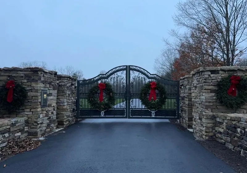 The Grand Estate Gate closed across a wet asphalt drive at dusk between broad dry-stacked stone piers, with a Christmas wreath and red bow hung on each leaf and on each pier.