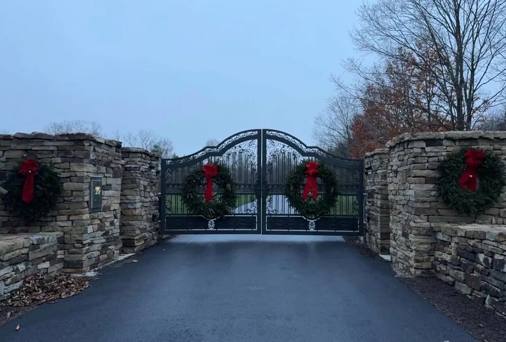 The Grand Estate Gate closed across a wet asphalt drive at dusk between broad dry-stacked stone piers, with a Christmas wreath and red bow hung on each leaf and on each pier.
