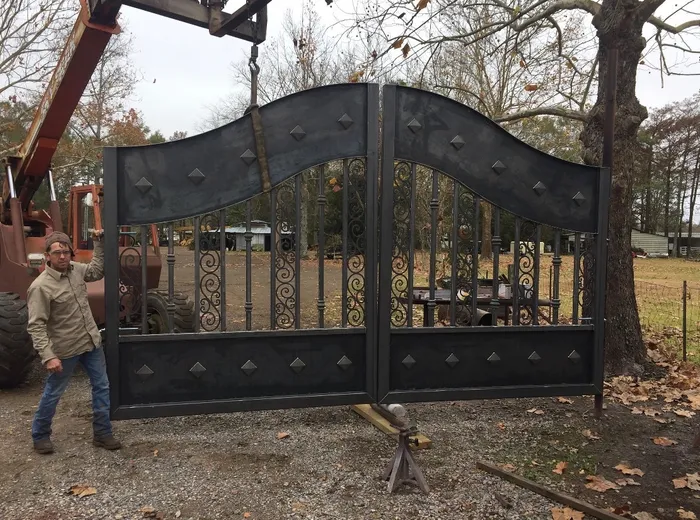 Both leaves of the Tampa Gate standing upright in a gravel yard before the finish coat, the plate still in bare dark steel and the scroll panels bright, slung by a strap from a tractor boom while a man steadies the left leaf.