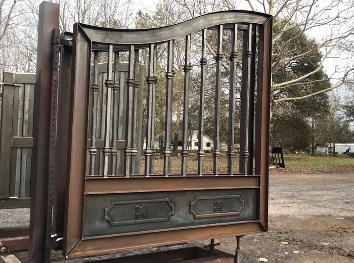 One leaf of the Fresina Gate on a stand in the yard in bare unpainted steel, its grey and brown surface showing the ogee rail, the collared balusters and the two rosette panels along the bottom.
