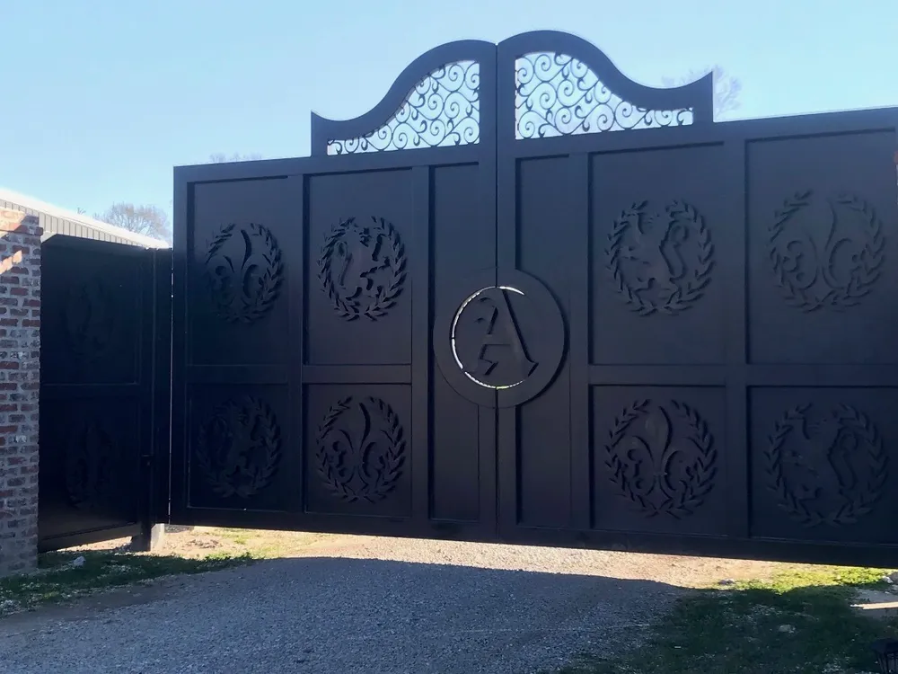 The Studio Gates closed across a gravel drive, wreath medallions standing off the plated panels and a scroll-filled arch rising over the centre, with a smaller matching gate open in the brick wall at left.