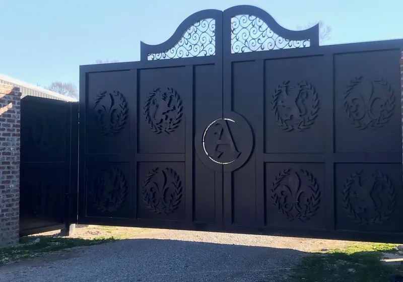 The Studio Gates closed across a gravel drive, wreath medallions standing off the plated panels and a scroll-filled arch rising over the centre, with a smaller matching gate open in the brick wall at left.