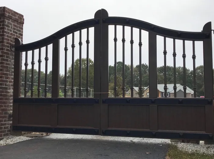 The gates square-on in a bronze-brown finish with near-black studded bands, the top rail curving up to the centre, hung from a brick column beside a gravel drive.