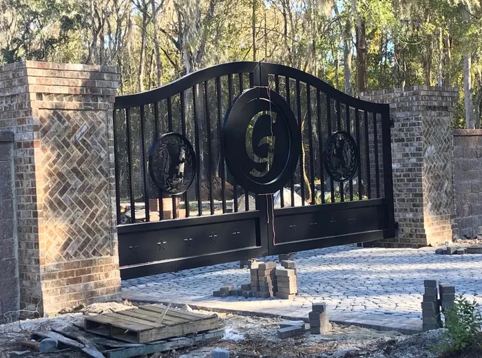 The lettered version of the gate closed between herringbone brick columns, low sun on the black steel, stacked pavers and a timber pallet still on the ground in front of it.