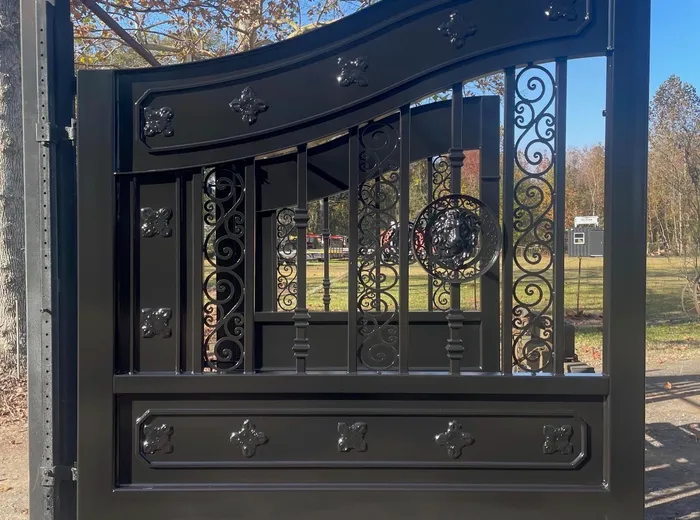 A single leaf of the New Yorker Gate propped upright on a concrete pad in high-gloss black, sunlight picking out the rosettes, the scroll panels and the lion's-head ring, with a tree in autumn colour above.
