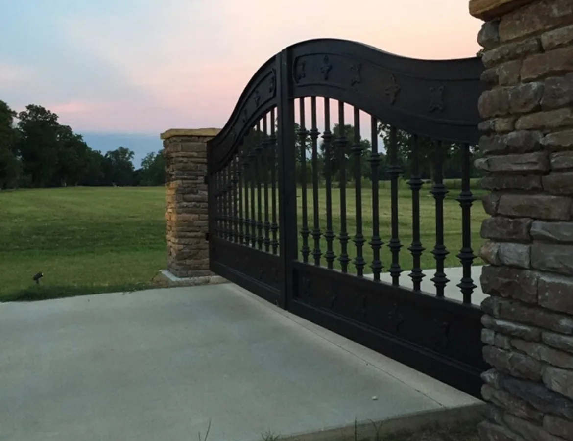 A J&L estate driveway gate closed at dusk between stacked-stone columns, its curved iron top rail against an orange sky.