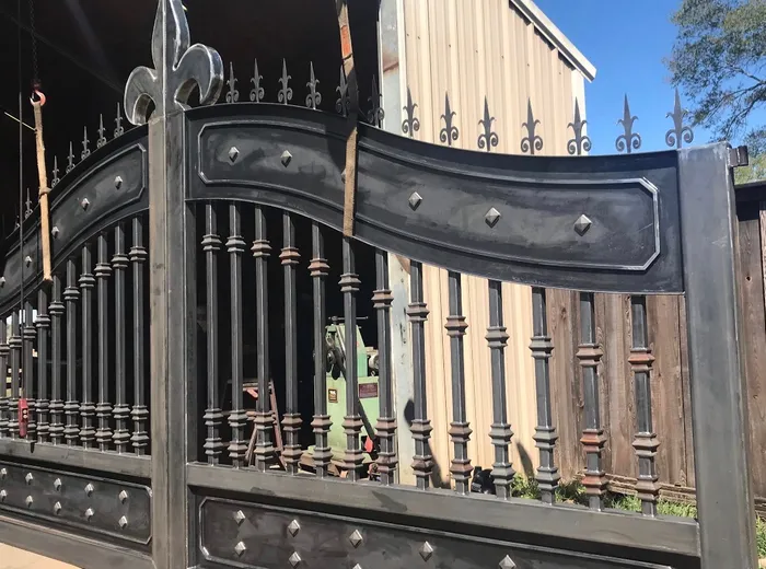 Both leaves in the shop yard before finishing, bare steel showing the curved top panel, the ringed balusters and the fleur-de-lis at the centre, with a lifting strap still attached.