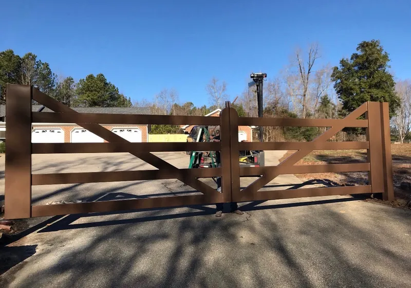 The Virginia Farm Gate closed square across a paved drive, its brown rails and raking diagonal braces set against a brick house with white garage doors and bare winter trees behind.
