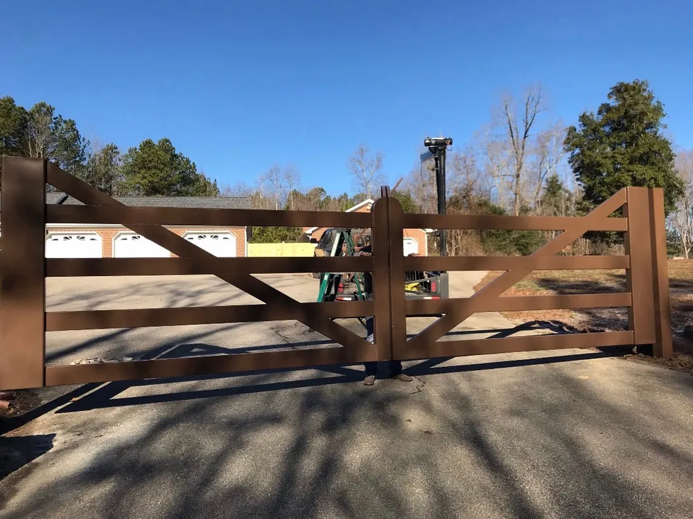 The Virginia Farm Gate closed square across a paved drive, its brown rails and raking diagonal braces set against a brick house with white garage doors and bare winter trees behind.