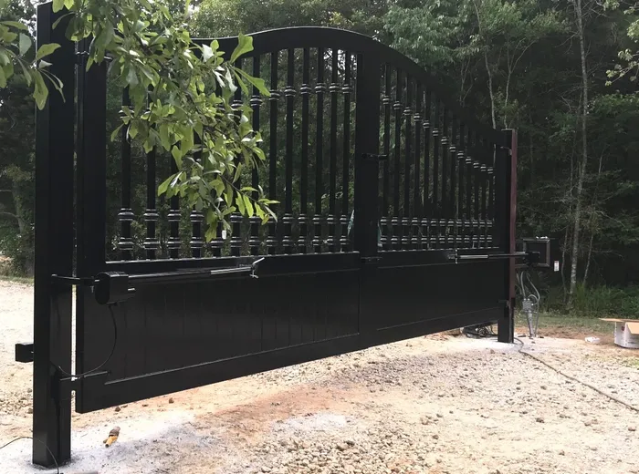 The Kling Gate closed on pale gravel with operator arms fitted along the inside of the mid rail and cabling dropping down the left post, a branch of leaves overhanging the left leaf.