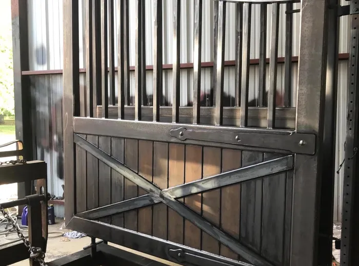 One leaf of the Stable Gate standing in the shop in bare steel, lifting eyes welded to the arched top rail and the X brace laid across the boarded lower panel.