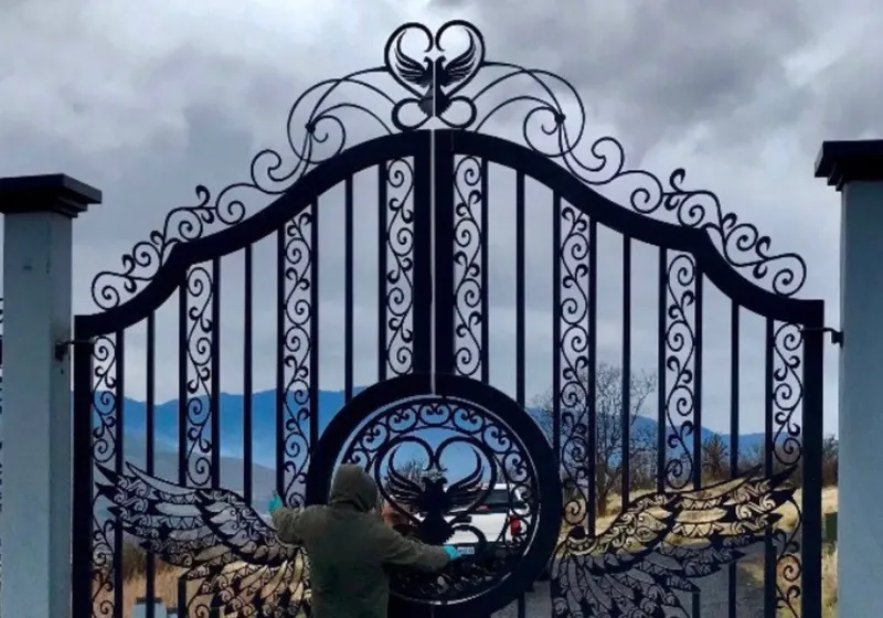 The Oregon Gate closed between pale masonry piers at the head of a gravel drive under heavy grey cloud, two people standing at the centre of the leaves, with blue ridgelines showing through the ironwork.