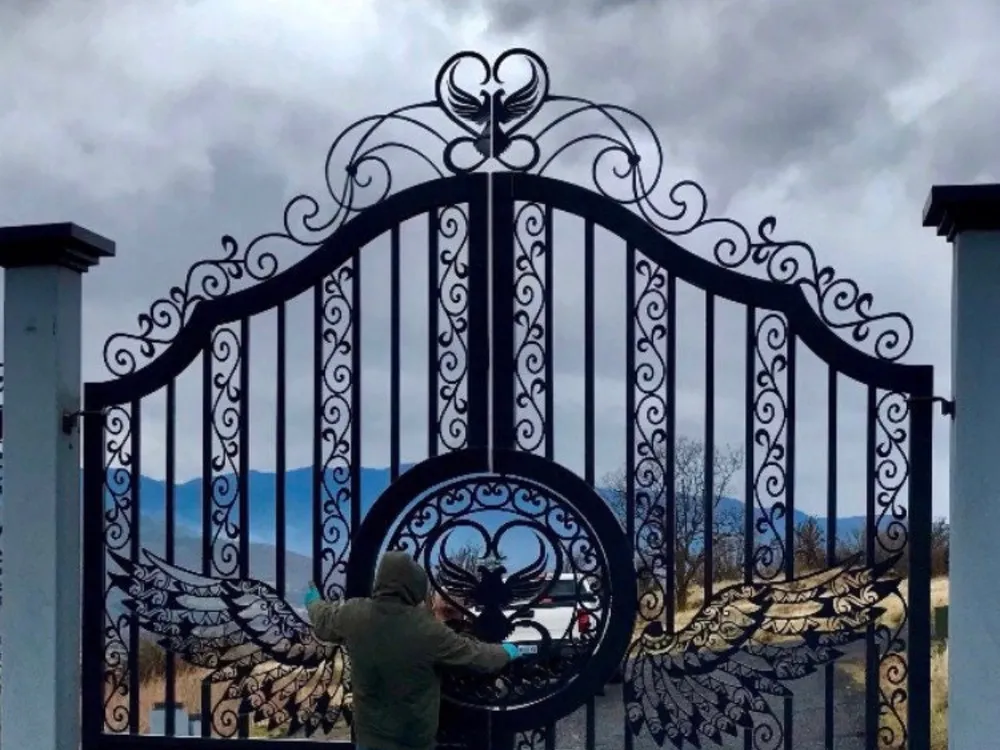 The Oregon Gate closed between pale masonry piers at the head of a gravel drive under heavy grey cloud, two people standing at the centre of the leaves, with blue ridgelines showing through the ironwork.