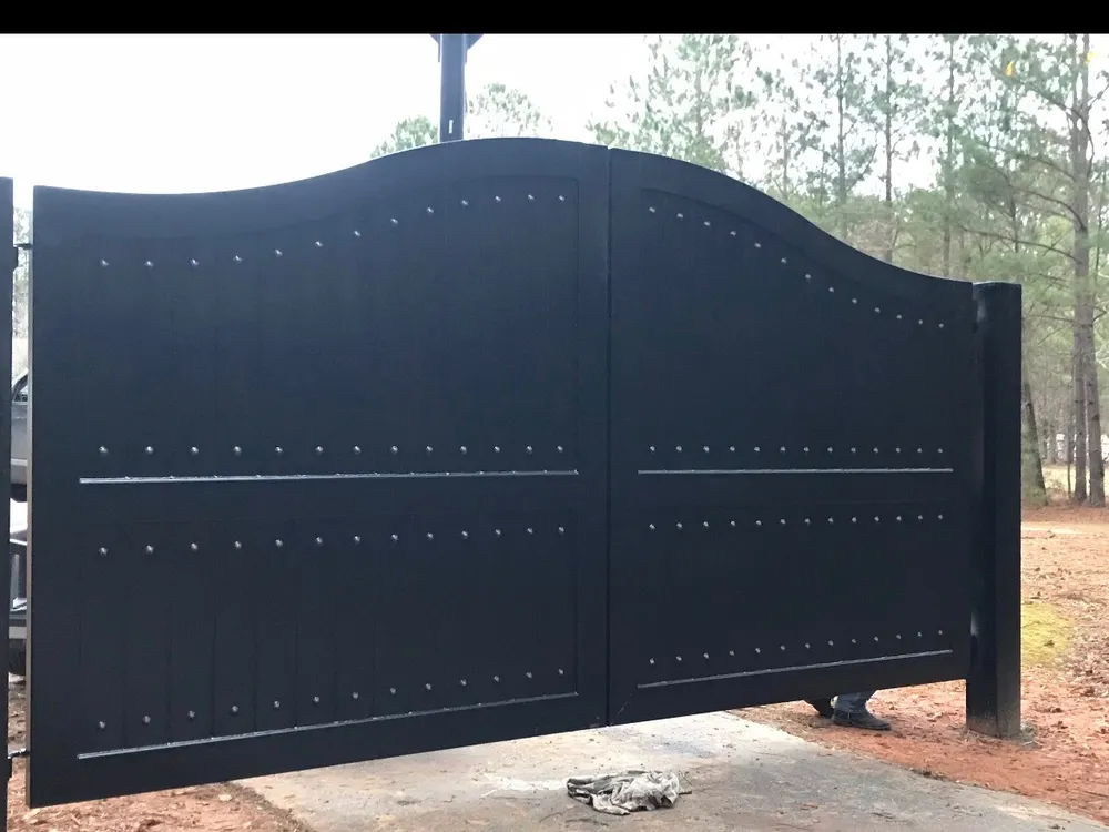 The Vigen Gate closed across a concrete apron in a stand of pines, its black boarded leaves lifting to a crest either side of the centre joint, the rows of domed studs picked out against the flat plate.