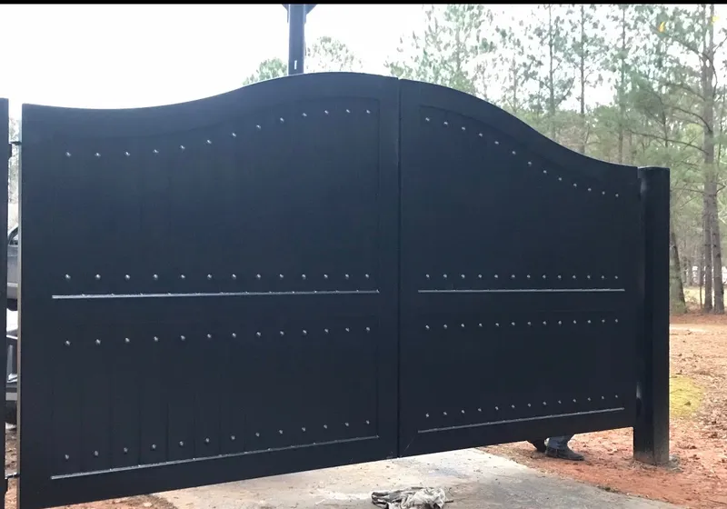 The Vigen Gate closed across a concrete apron in a stand of pines, its black boarded leaves lifting to a crest either side of the centre joint, the rows of domed studs picked out against the flat plate.