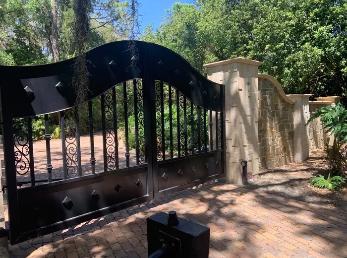 The Tampa Gate seen along the paver drive at an angle, the wave of the top rail and its pyramid studs catching the light beside the stone column.