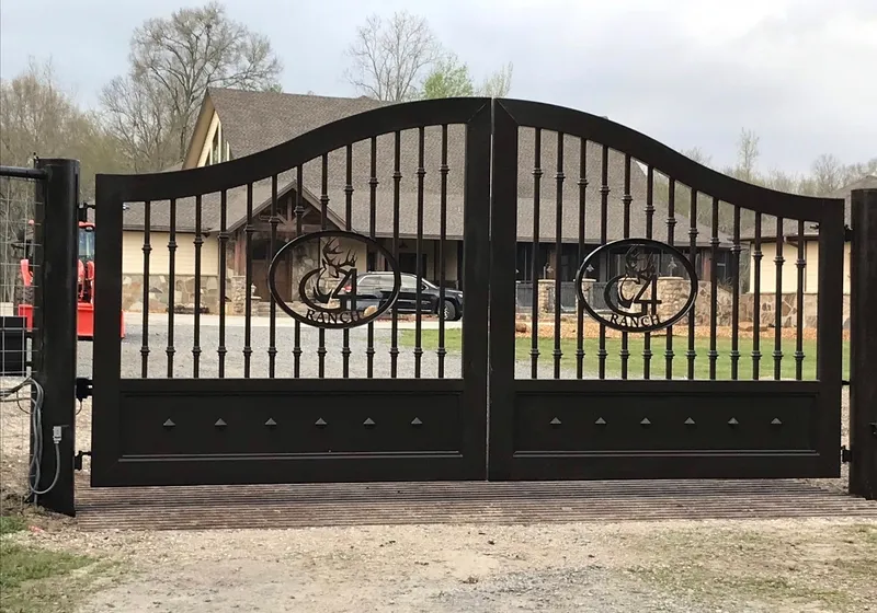 The Cali Gate closed square over a slatted cattle guard at the head of a gravel drive, its arched black leaves and two cut medallions standing in front of a cream-sided house with a stone base and bare-limbed trees behind.