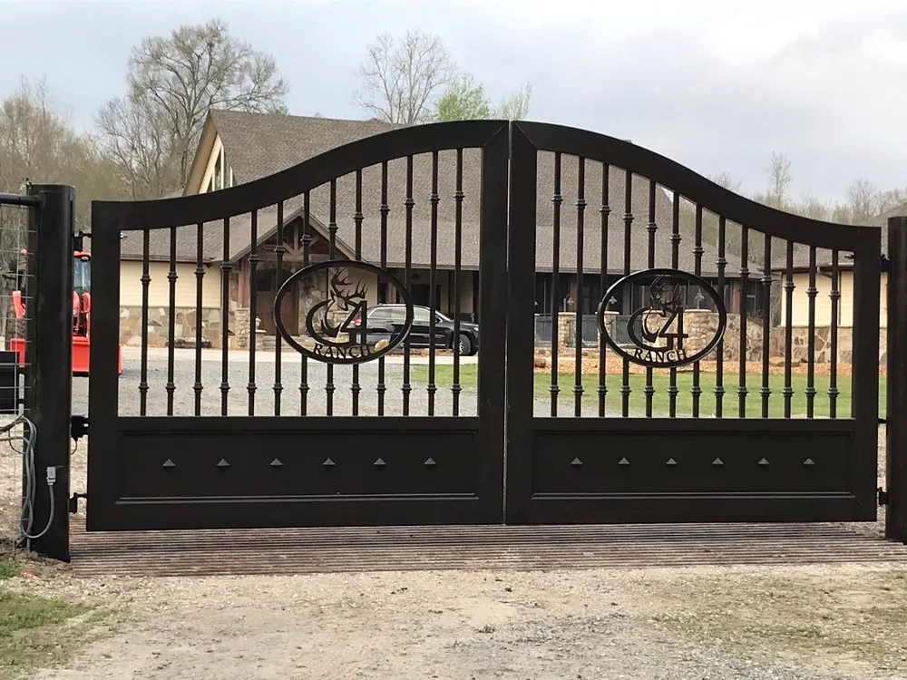 The Cali Gate closed square over a slatted cattle guard at the head of a gravel drive, its arched black leaves and two cut medallions standing in front of a cream-sided house with a stone base and bare-limbed trees behind.