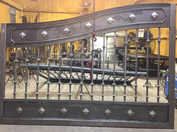 A single leaf standing inside the shop under the hoist chain, its studded top rail, turned balusters and studded bottom panel all in bare steel.