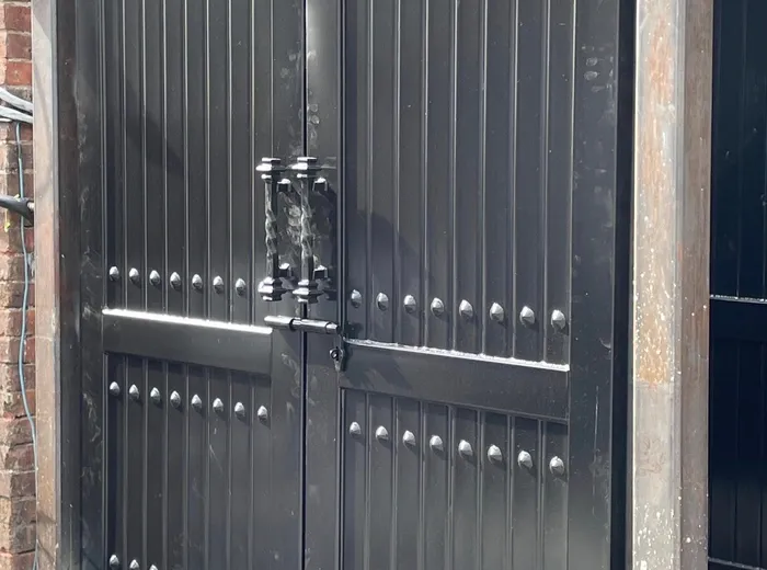 A narrower square-topped pair in glossy black, deeply ribbed and heavily studded, hung on steel posts in a brick opening with a twisted-bar pull at the meeting stiles.