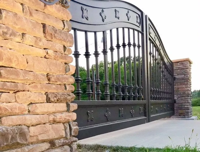 Close view of the gate where it meets its stacked-stone column, the rosette-set lower band at eye level and the turned balusters running away to the far column.